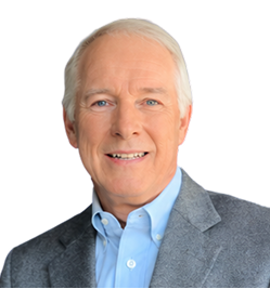 Portrait of a smiling older man with white hair wearing a gray blazer and light blue collared shirt against a white background.