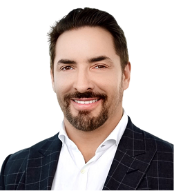 Headshot of a man with dark hair, a beard, and a mustache, wearing a white shirt and dark checkered suit jacket, smiling against a white background.