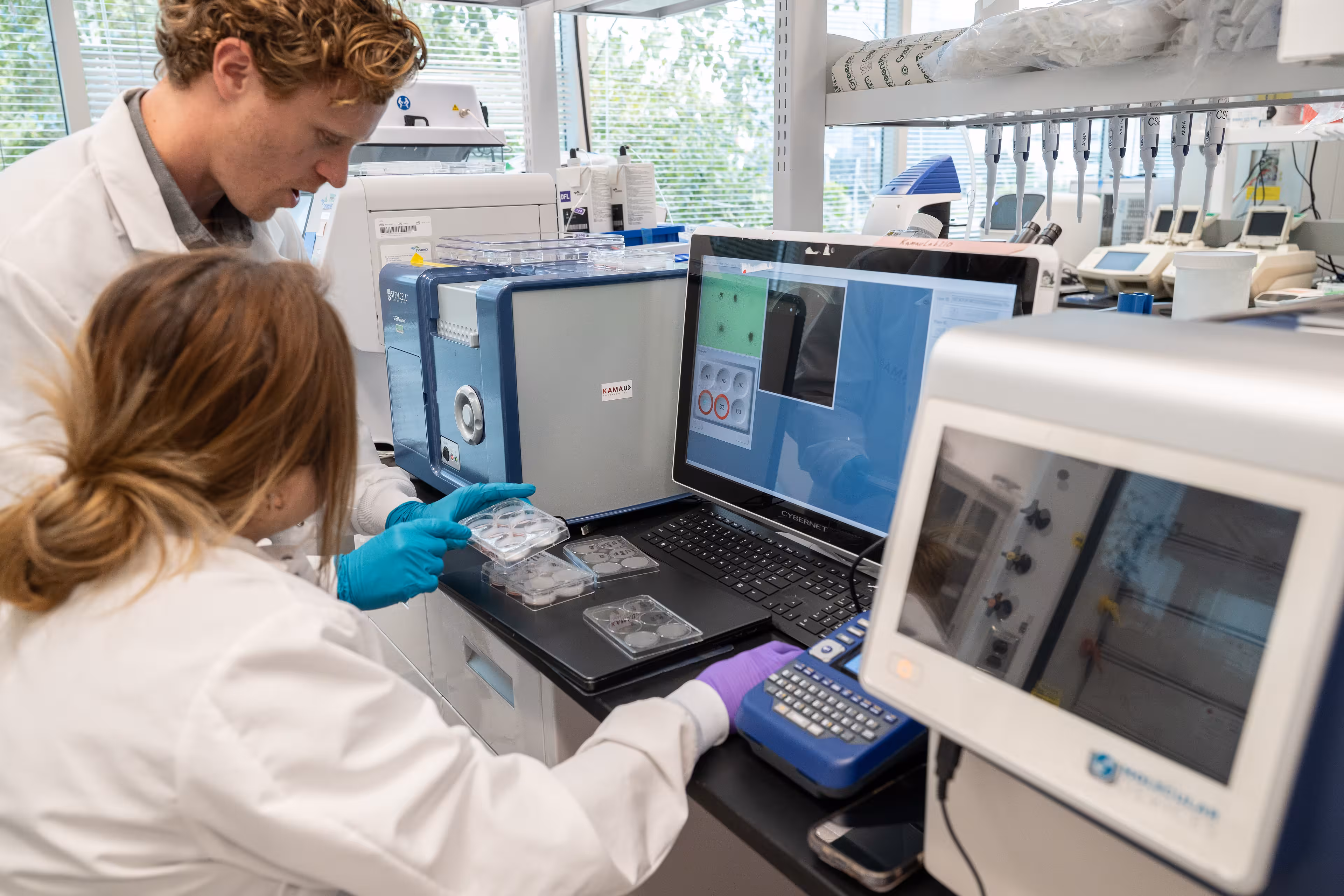 Two scientists in white lab coats working in a laboratory surrounded by scientific equipment, with one holding petri dishes and the other operating a device connected to a laptop displaying data.
