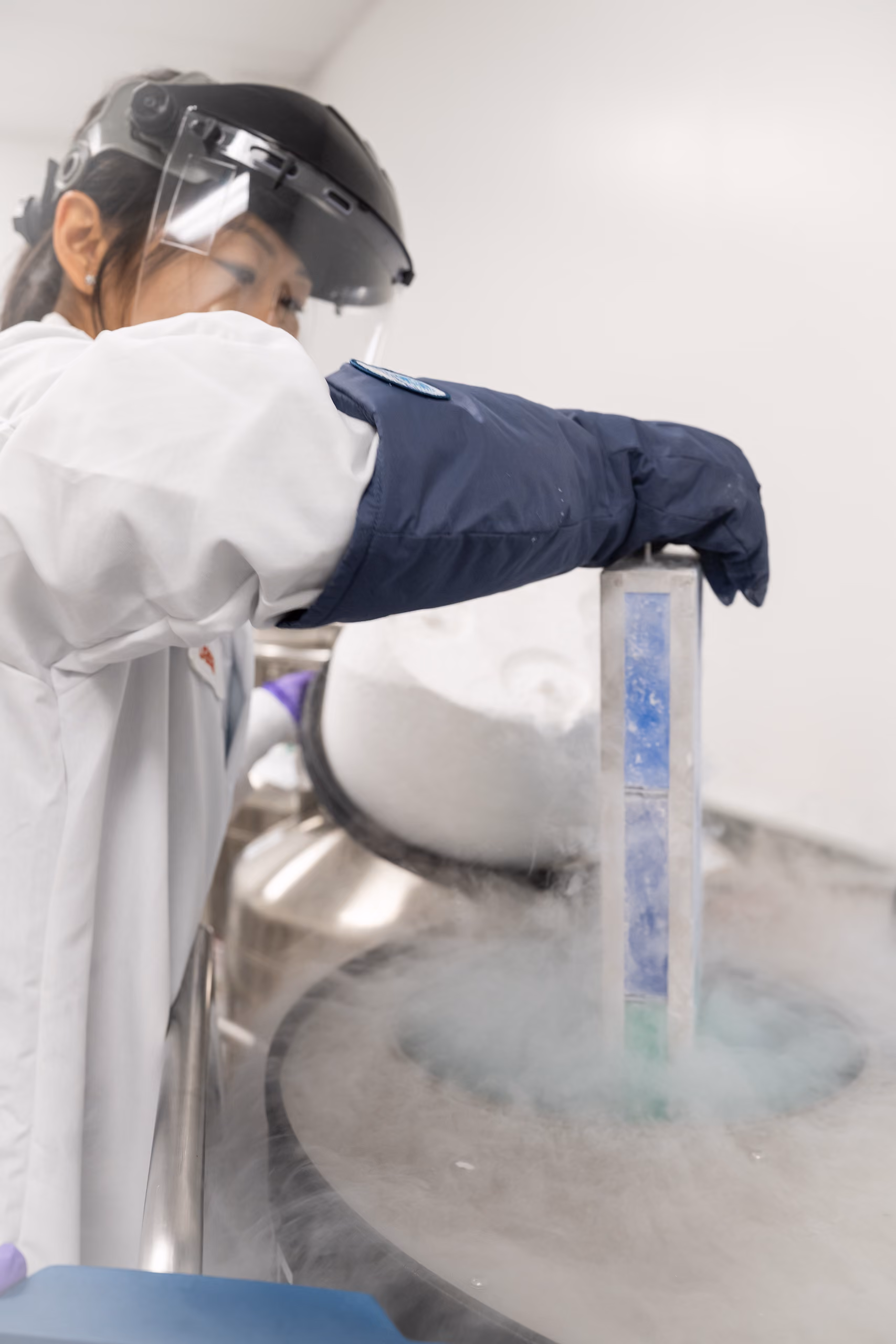A laboratory technician wearing a white coat, face shield, and thick protective glove is holding a container above a large metal tank emitting vapor, likely involved in cryogenic or cold storage work.