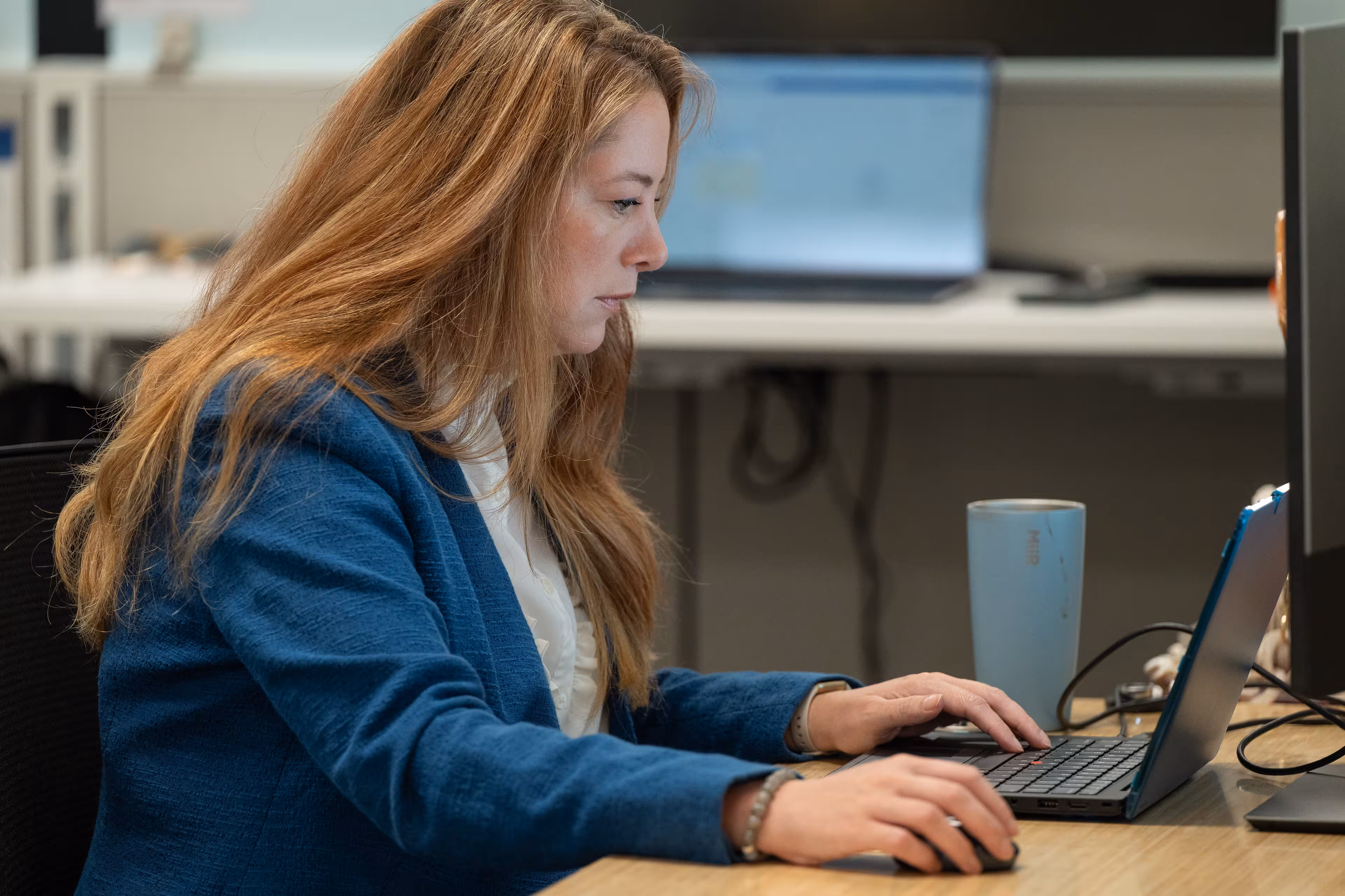 Woman with long blonde hair wearing a blue blazer, sitting at a desk and working on a small laptop with an external mouse, with a blue tumbler and a desktop monitor nearby.