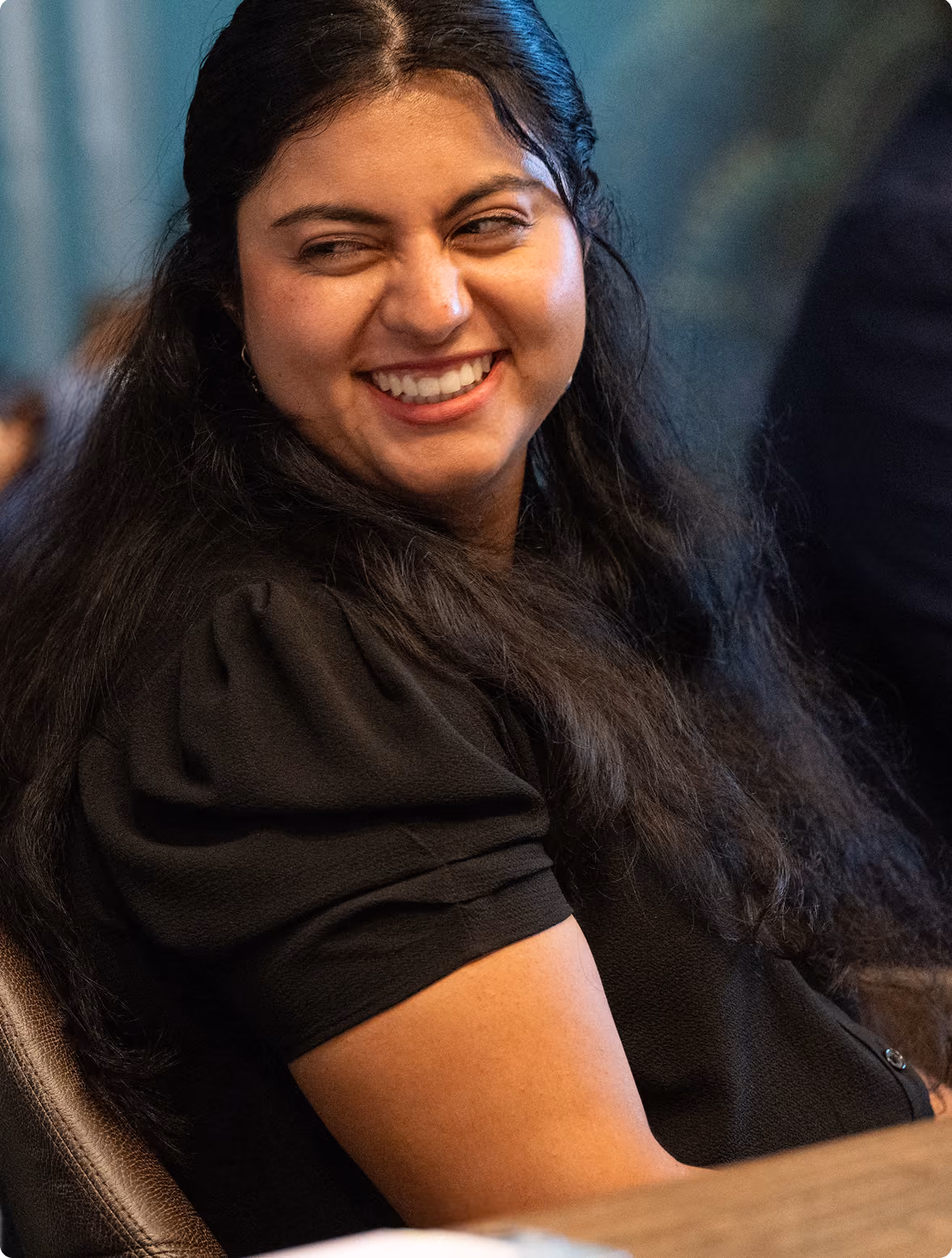 Woman with long dark hair wearing a black short-sleeve top, smiling and looking to the side in an indoor setting.