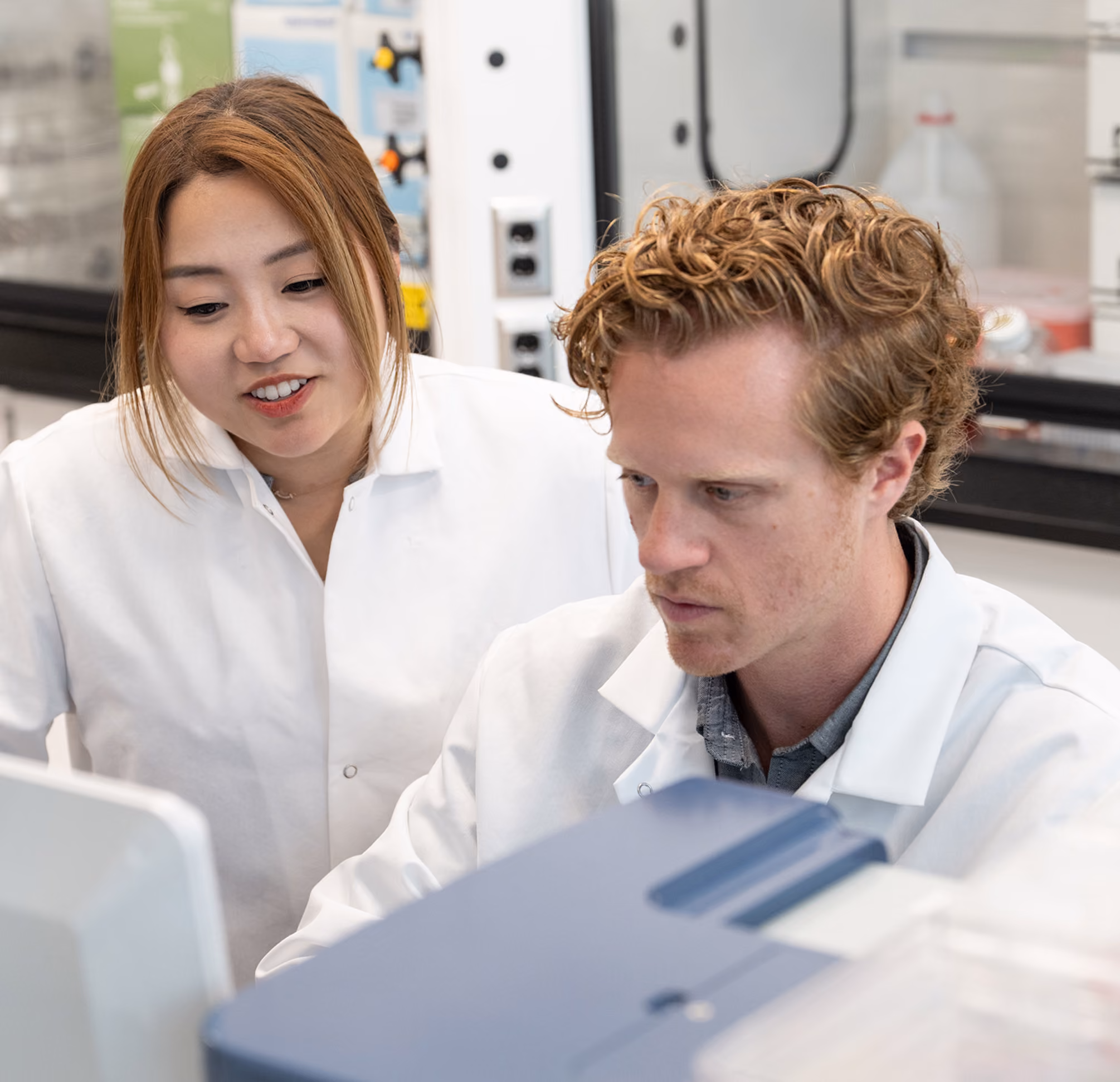 Two scientists wearing white lab coats working together in a laboratory, looking intently at a computer screen.