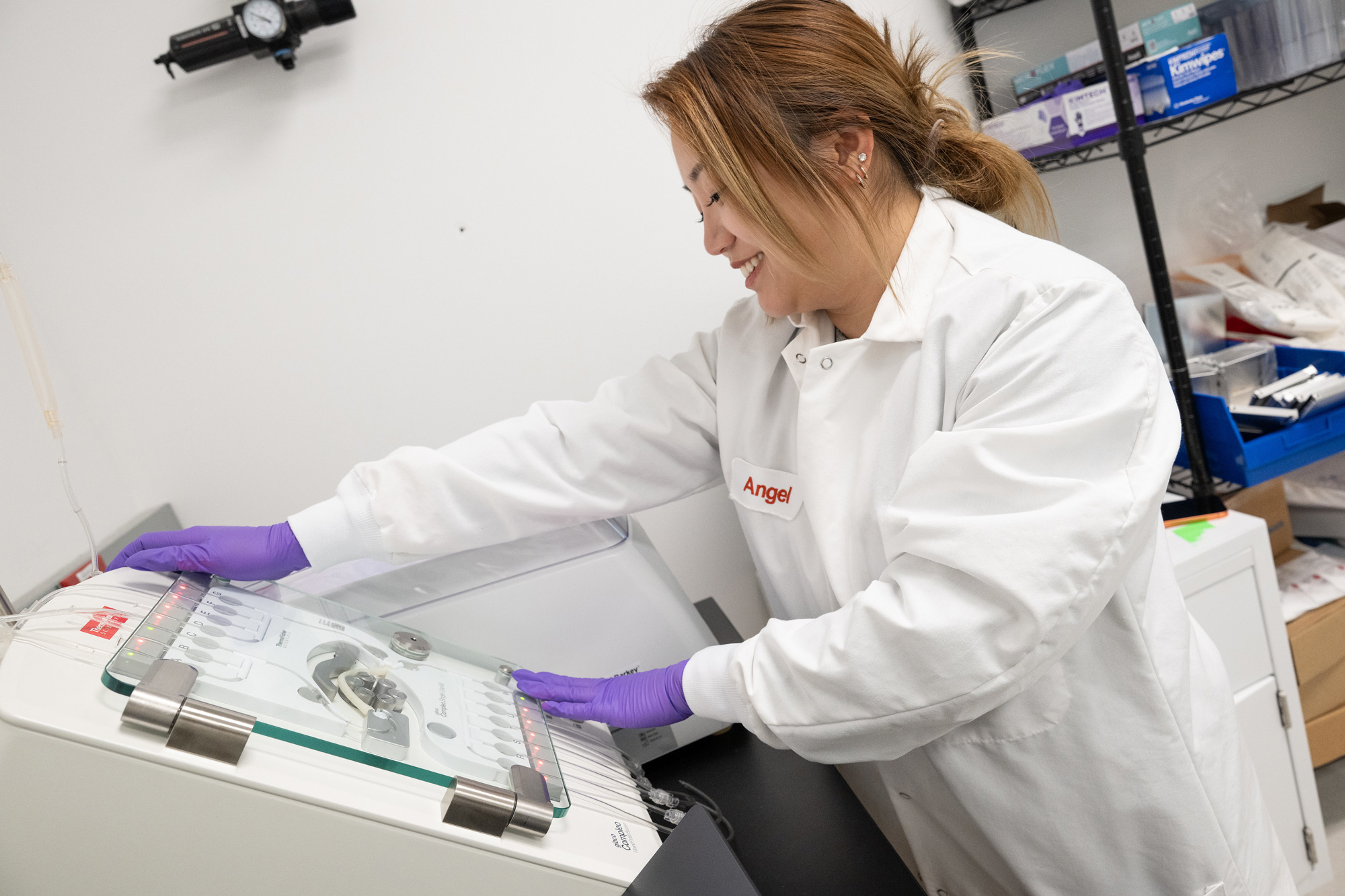 A smiling lab technician wearing a white coat and purple gloves operates a lab machine with tubes and blinking red lights in a laboratory setting.