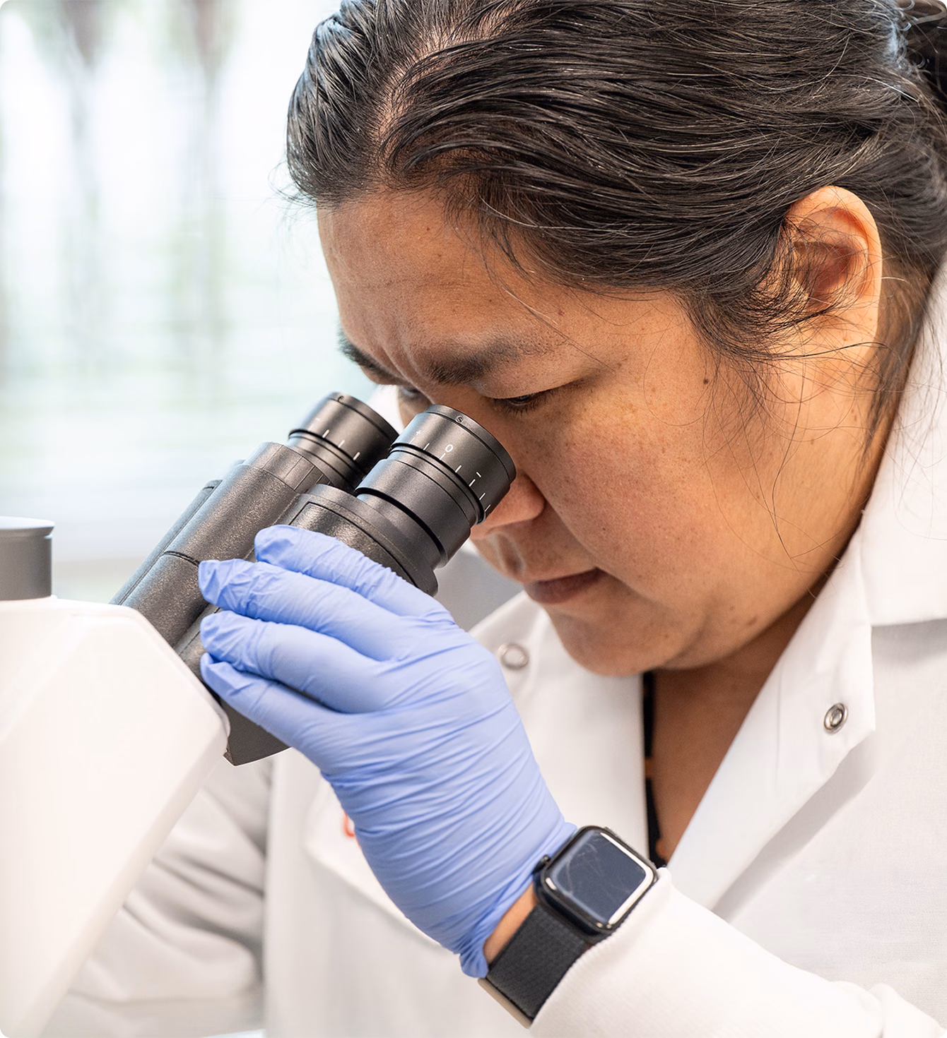 Scientist wearing a white lab coat and blue gloves closely examining a sample through the eyepieces of a microscope in a laboratory setting.