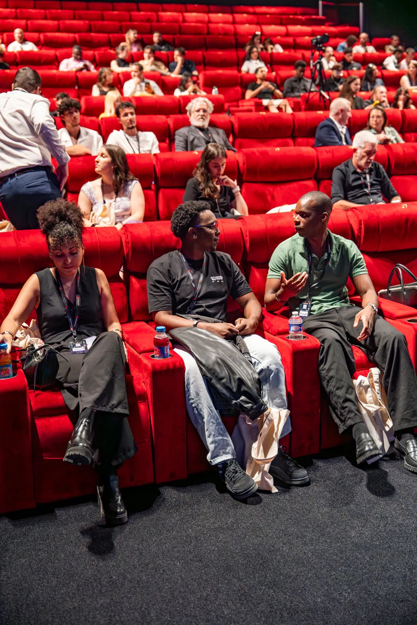 Public assis dans la salle de cinéma rouge, discussion animée avant l'événement Accélération.