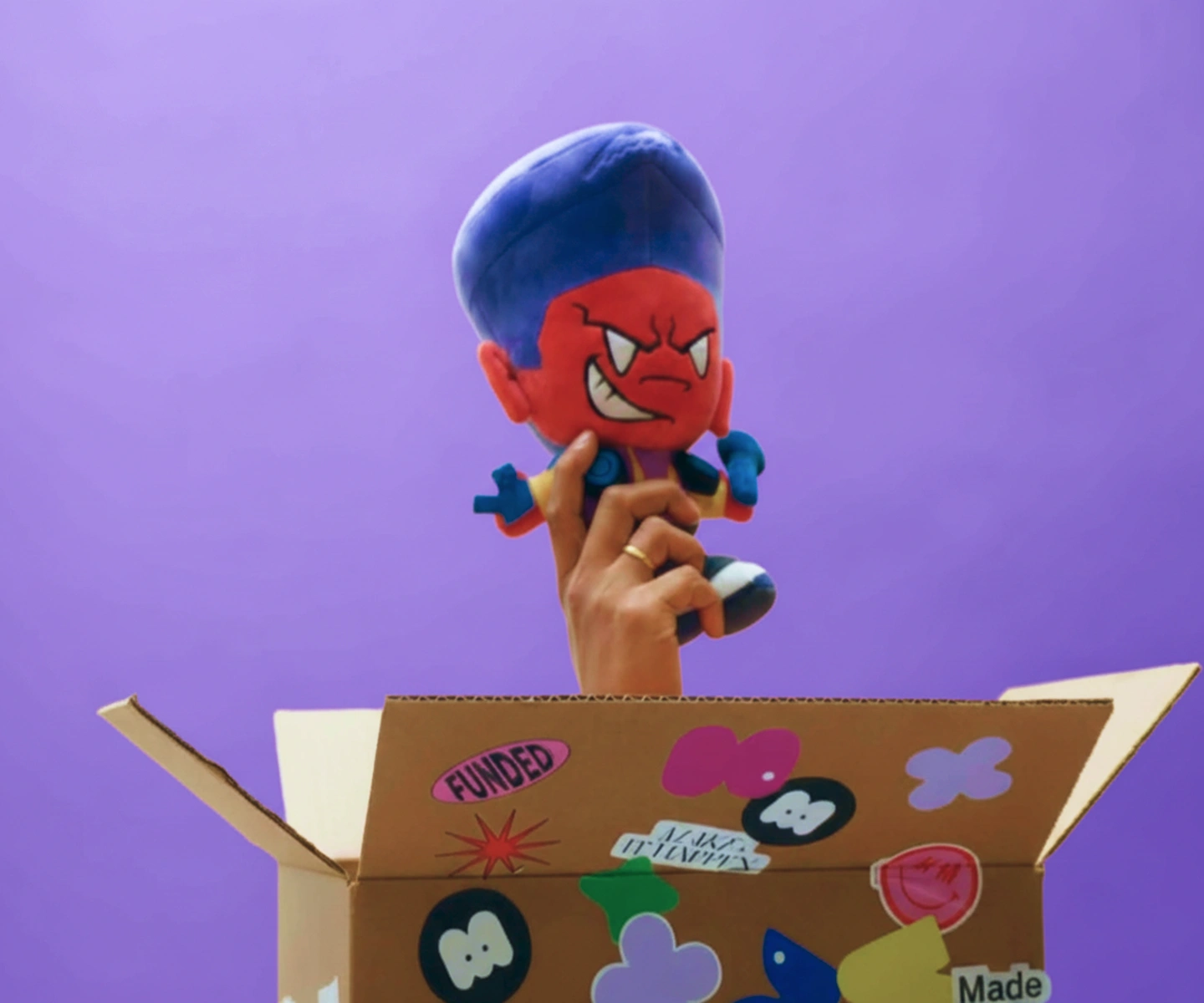 A hand with a gold ring holding a red-faced plush toy with blue hair and an angry expression above an open cardboard box decorated with colorful stickers against a purple background.