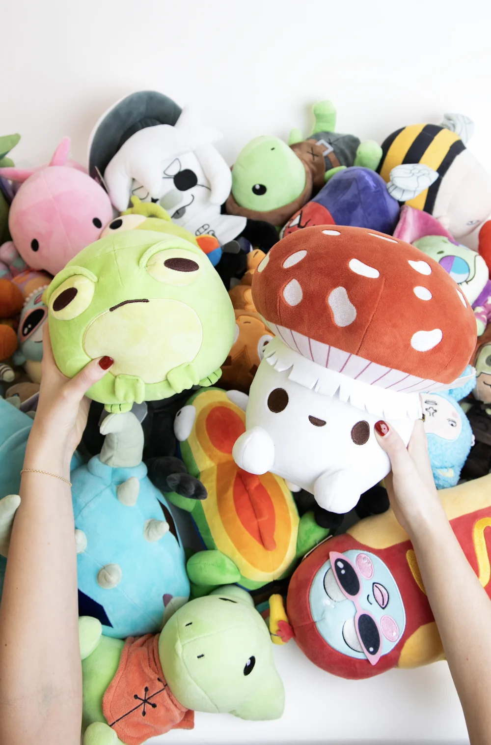 A collection of colorful plush toys including a large green frog held by a left hand and a white mushroom with a red spotted cap held by a right hand, surrounded by other various stuffed animals and characters on a white surface.