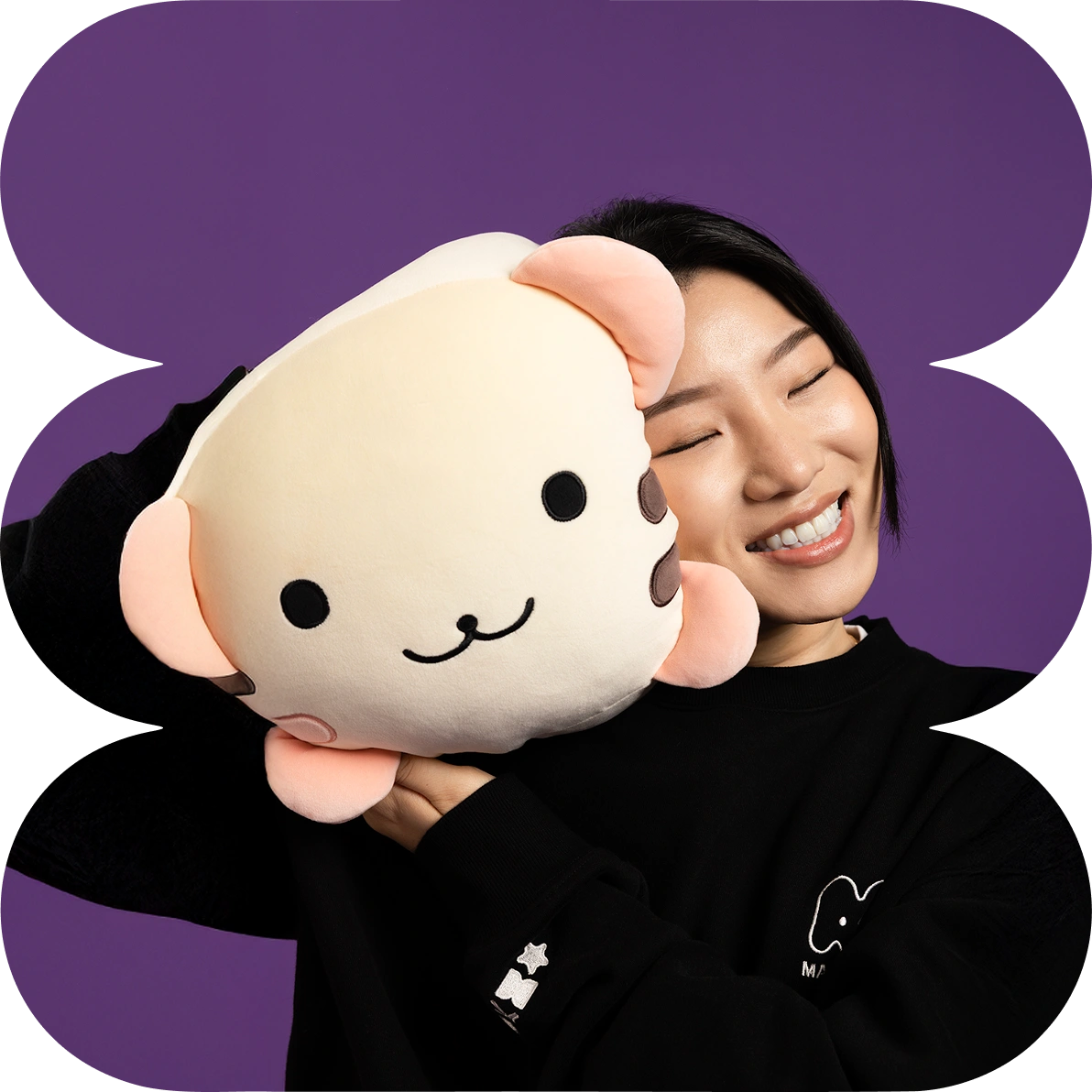 Smiling woman with eyes closed holding a large round beige stuffed animal with a cute face and pink ears against a purple background.