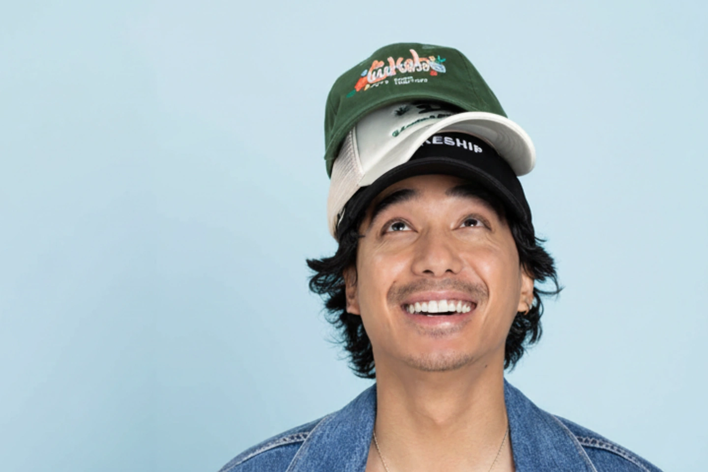 Smiling man with curly dark hair wearing a denim jacket and three stacked caps—black, white, and green with colorful embroidery—on his head, looking upward against a light blue background.