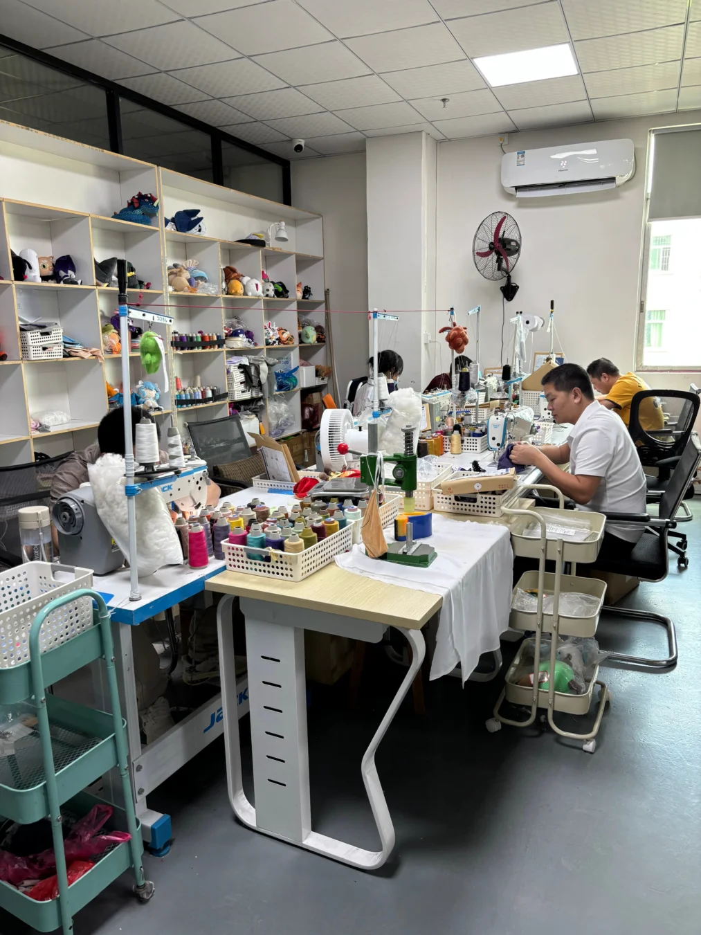 A sewing workshop with multiple workers seated at sewing machines at a long table, surrounded by shelves holding various stuffed toys and spools of thread, with fans and an air conditioner mounted on the walls.
