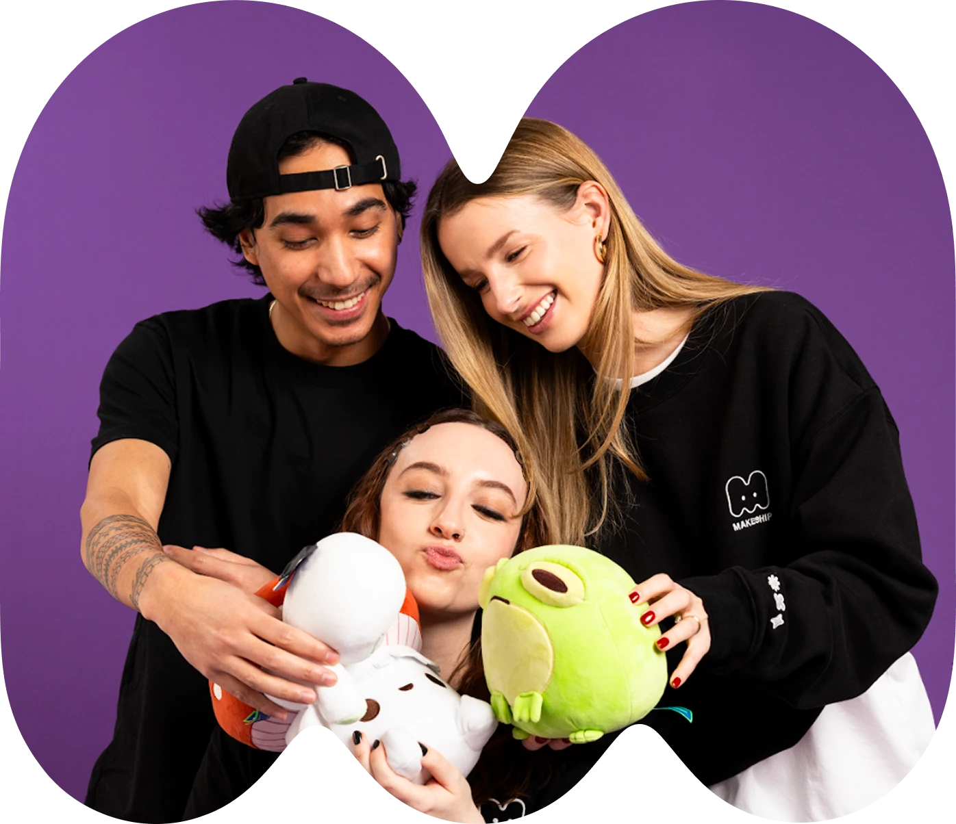 Three young people smiling and holding plush toys, with a purple background. The person on the left wears a black cap and t-shirt, the person in the center puckers lips, and the person on the right in a black sweatshirt leans in.