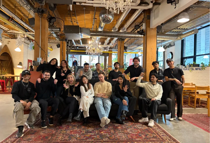 A diverse group of 15 people posing and smiling together in a cozy, industrial-style room with exposed pipes, wooden beams, chandeliers, and large windows.