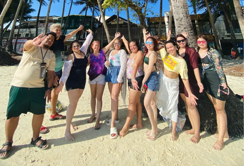 Group of nine people standing on a sunny sandy beach with palm trees and buildings in the background, smiling and waving at the camera.