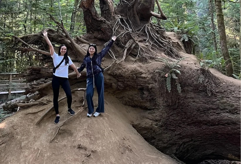 Two women standing and smiling on the large exposed root system of an uprooted tree in a forest, both with one arm raised in a joyful gesture.