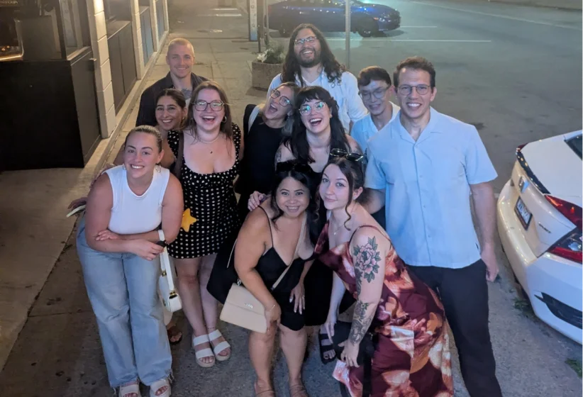 Group of eleven people posing closely together on a sidewalk at night, smiling and looking up at the camera, with a parked white car visible on the right side.