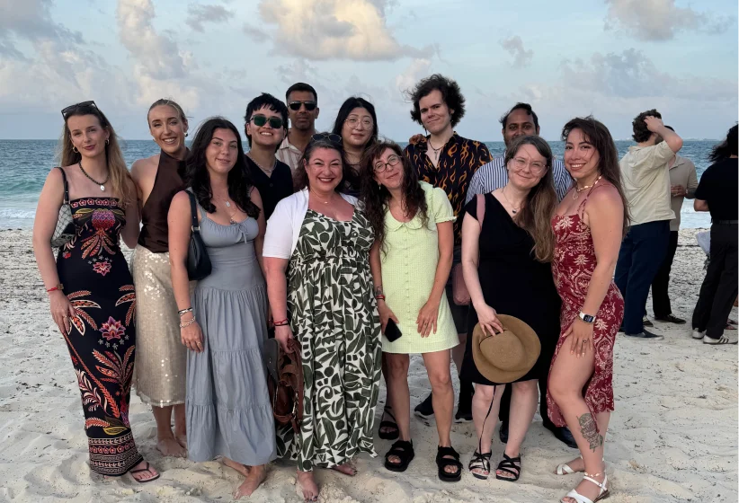 Group of twelve diverse adults standing on a sandy beach near the ocean during late afternoon, smiling and posing for a photo under a cloudy sky.