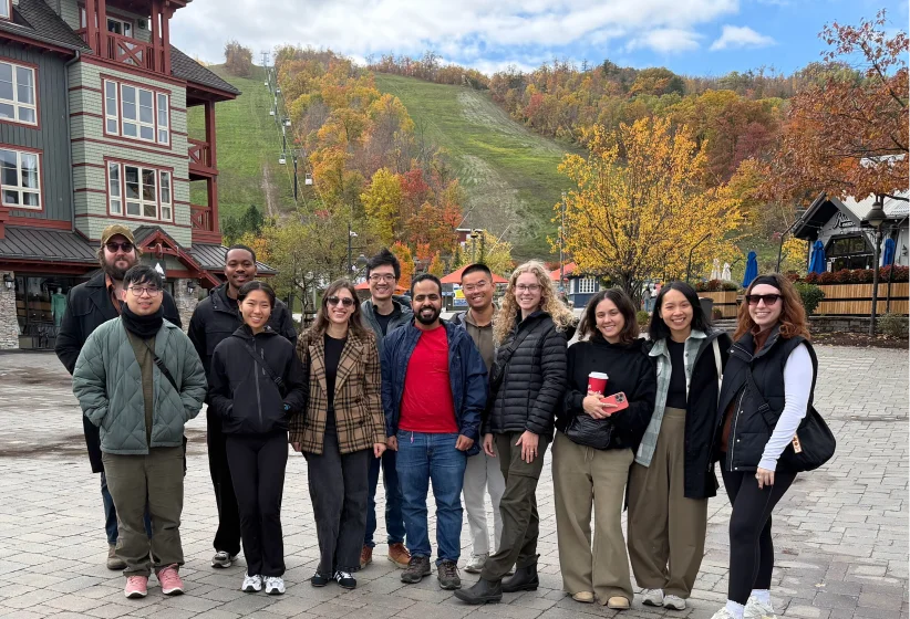 Group of twelve people standing together outdoors on a paved area with fall-colored trees and a ski slope in the background, dressed in casual jackets and pants.