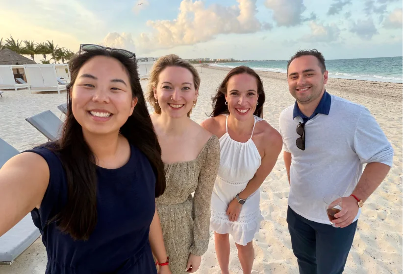 Four smiling adults posing for a selfie on a sandy beach near the ocean with partly cloudy sky and palm trees in the background.
