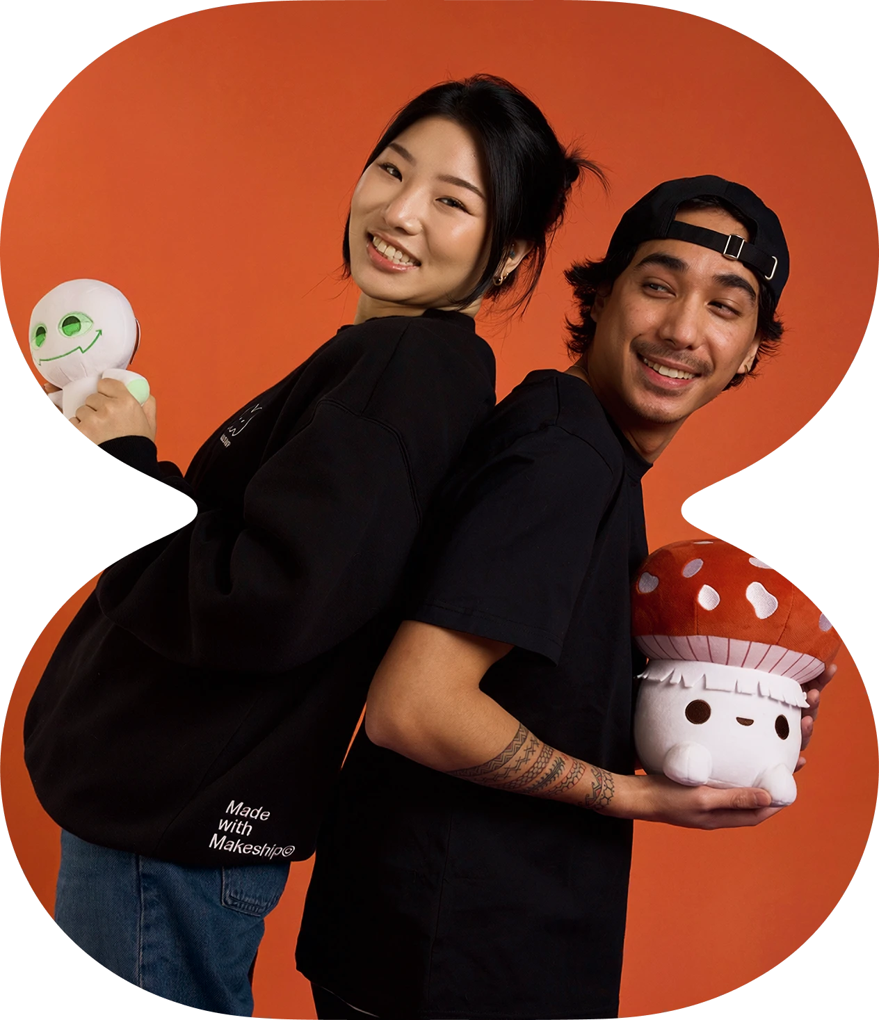 A woman and a man stand back to back against an orange background, both smiling and holding plush toys; the woman holds a green-eyed white plush and the man holds a mushroom-shaped plush. The woman wears a black sweatshirt with 'Made with Makeship' on the lower side and blue jeans, while the man wears a black t-shirt and a backward black cap, showing a tattoo on his forearm.