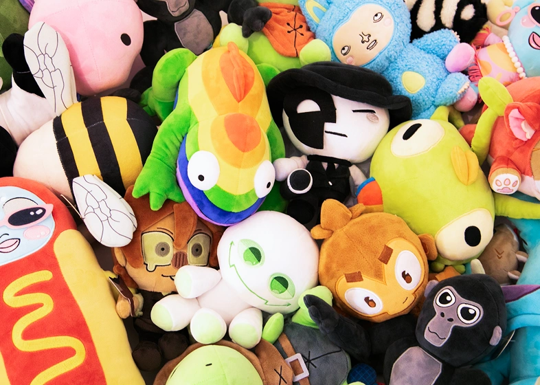 A colorful assortment of various plush toys closely packed together, including a multicolored frog, a hot dog with a smiling face, a white ghost-like character with green eyes, a black and white toy wearing a hat, and other assorted animal and fantasy plushies.