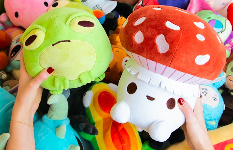 Hands holding two plush toys: a green frog with a tired expression on the left and a white mushroom character with a red cap with white spots on the right, surrounded by other colorful plush toys.