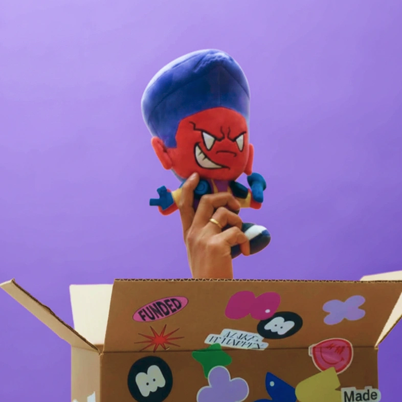 Hand holding a red-faced cartoon plush toy with blue hair and pointed white teeth above a decorated cardboard box against a purple background.