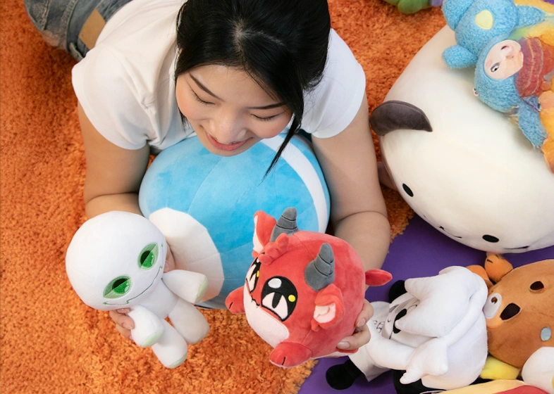 Young woman lying on an orange carpet, smiling and holding two plush toys, one white with big green eyes and one red with horns, surrounded by other colorful stuffed animals.