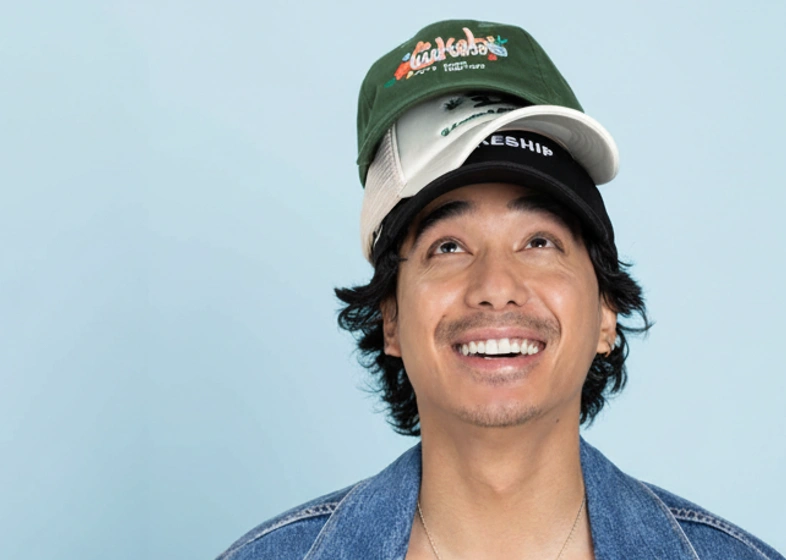 Smiling man with dark hair and mustache wearing three stacked caps in green, beige, and black colors, looking upward against a light blue background.