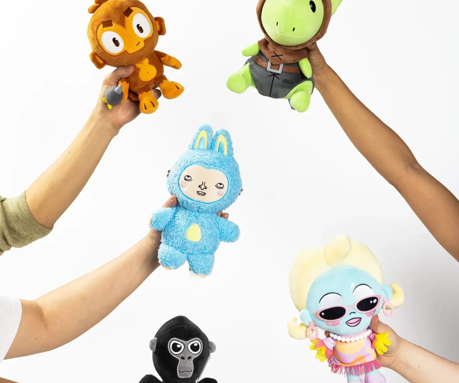 Five colorful plush toys held up by various hands against a white background, including an orange monkey, a green turtle in a brown hood, a blue bunny-like character with a human face, a black gorilla, and a blue-haired doll with sunglasses and pink earrings.