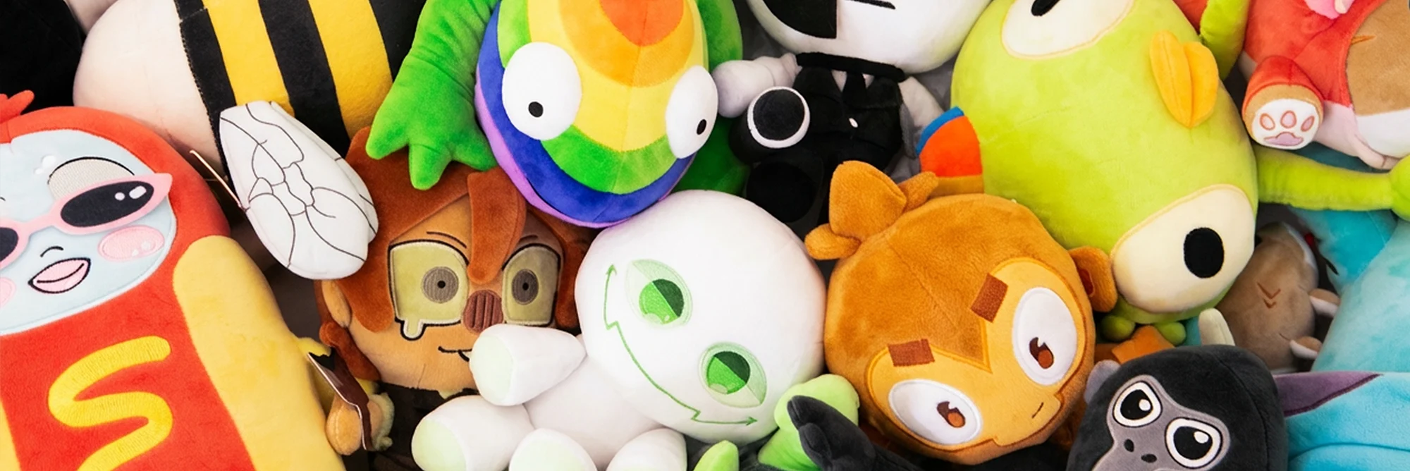 A colorful pile of assorted plush toys featuring various cartoon characters, including a red character with sunglasses, a white smiling creature with green eyes, a green frog with big eyes, a yellow bee, and other vibrant stuffed animals.