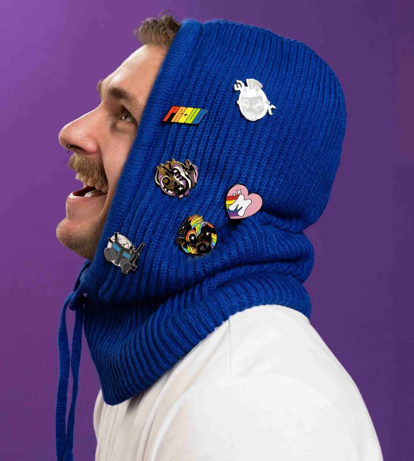 Side profile of a smiling person wearing a blue knitted hood adorned with six colorful pins, set against a purple background.
