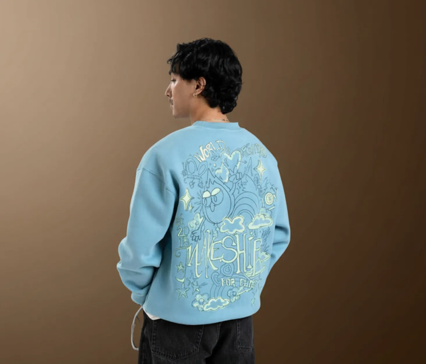 Man with short black curly hair wearing a light blue sweatshirt with a colorful cartoon-style design and text on the back, standing against a brown gradient background, facing sideways away from the camera.