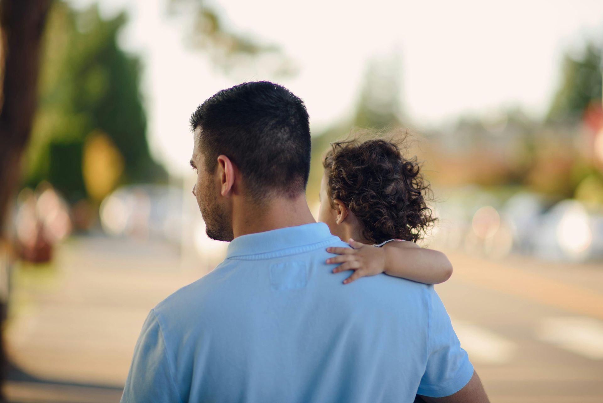 Adult carrying a small child on their shoulder on a sunlit street, both seen from behind
