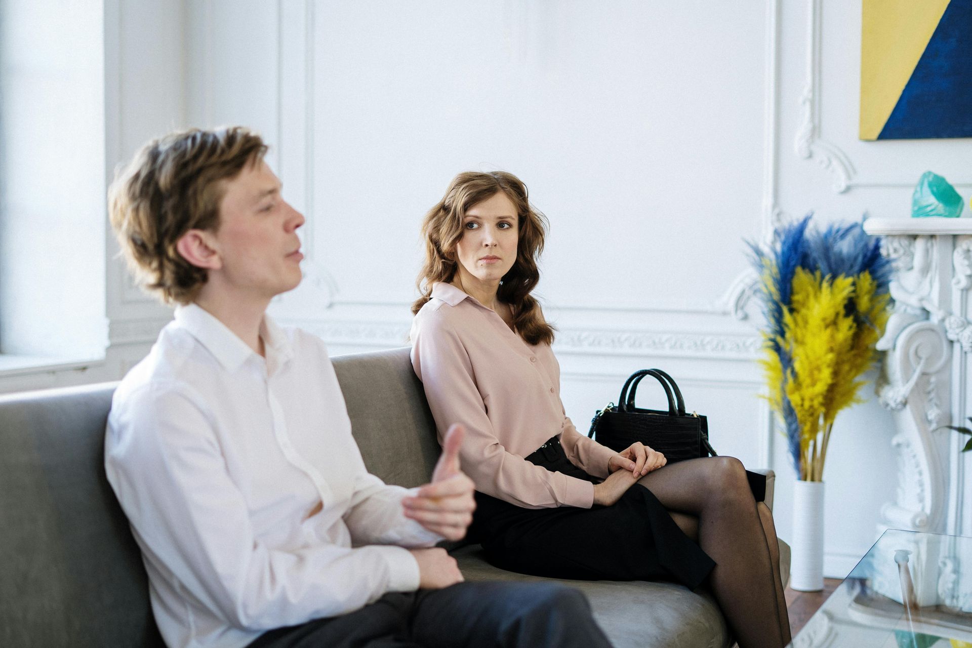 Two people sit on a couch in a bright room, one holding a black handbag near a vase of yellow and blue flowers.