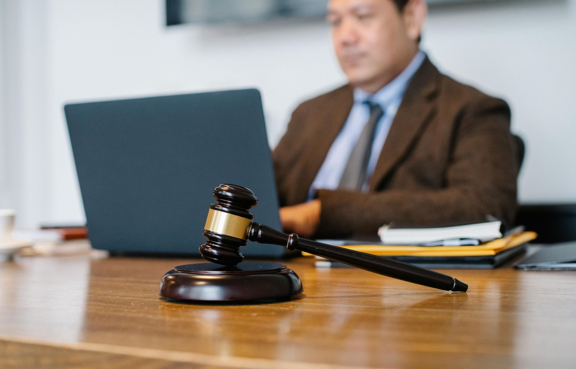 Judge’s gavel on a desk in front of a blurred lawyer working on a laptop in a courtroom setting
