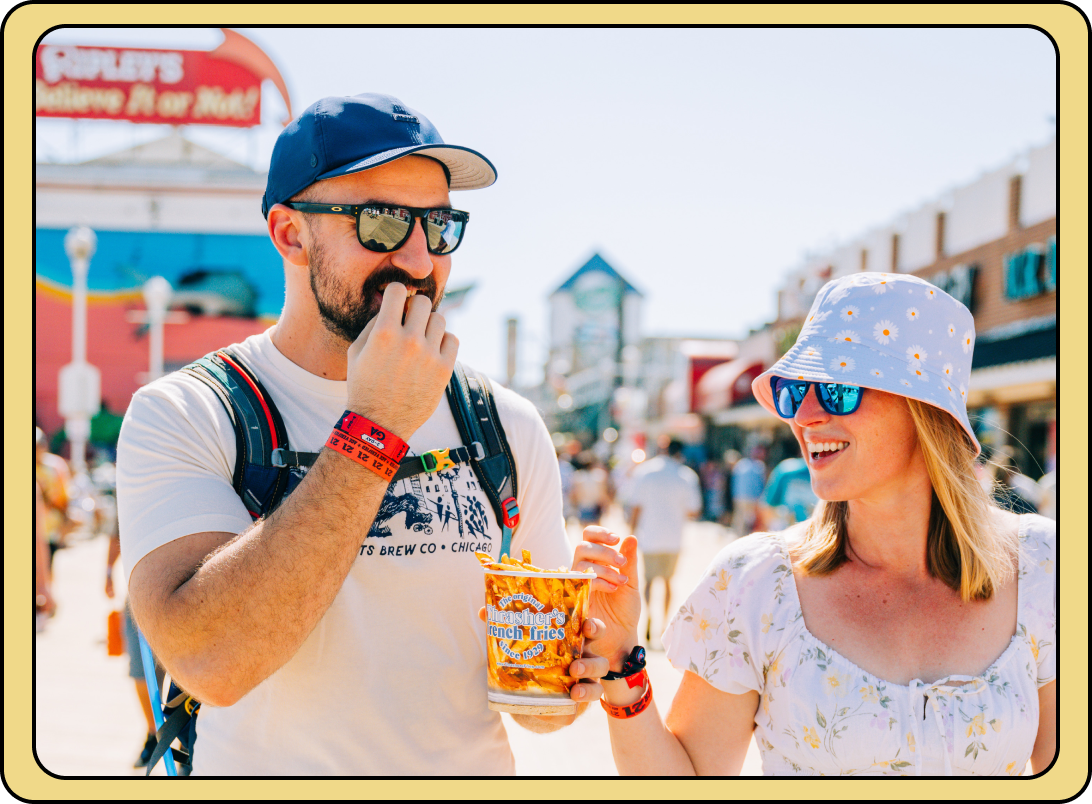 Two festivalgoers in sunglasses and wristbands sharing boardwalk fries on a sunny boardwalk