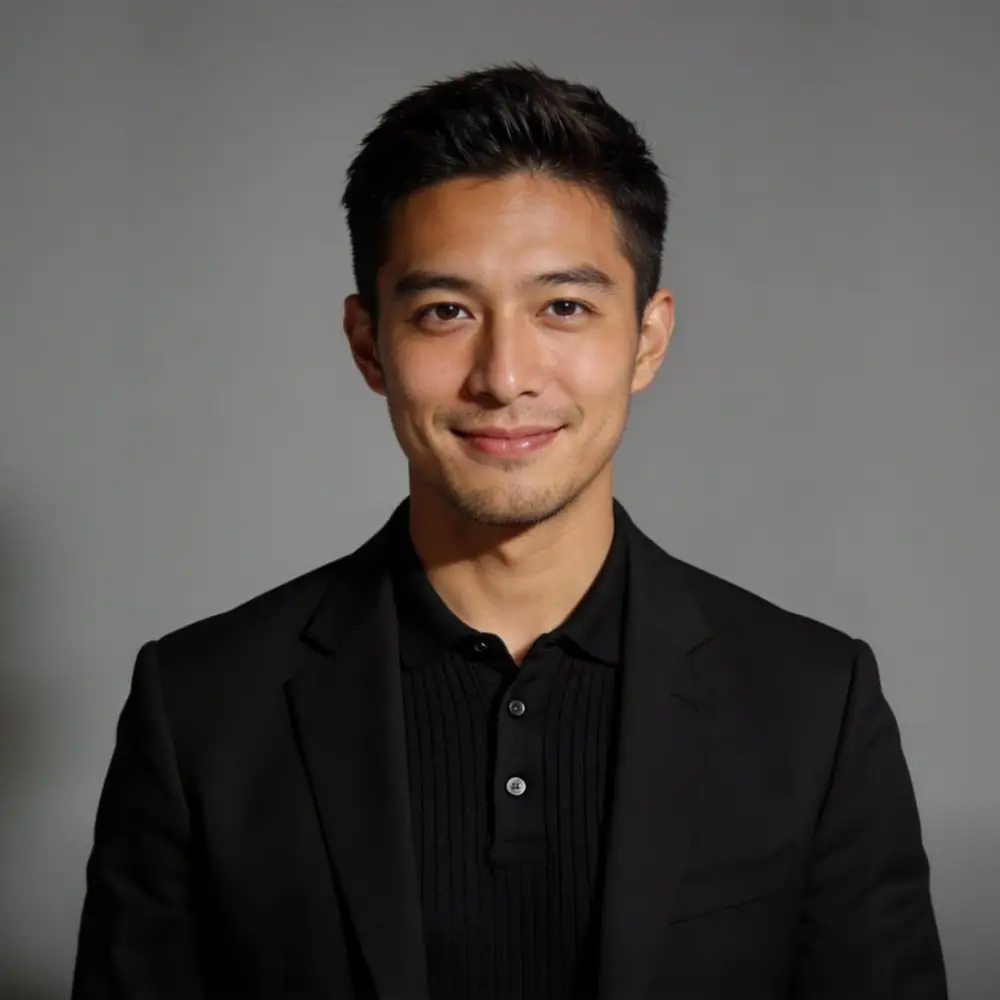 Smiling young man with short black hair wearing a black blazer and shirt against gray background.