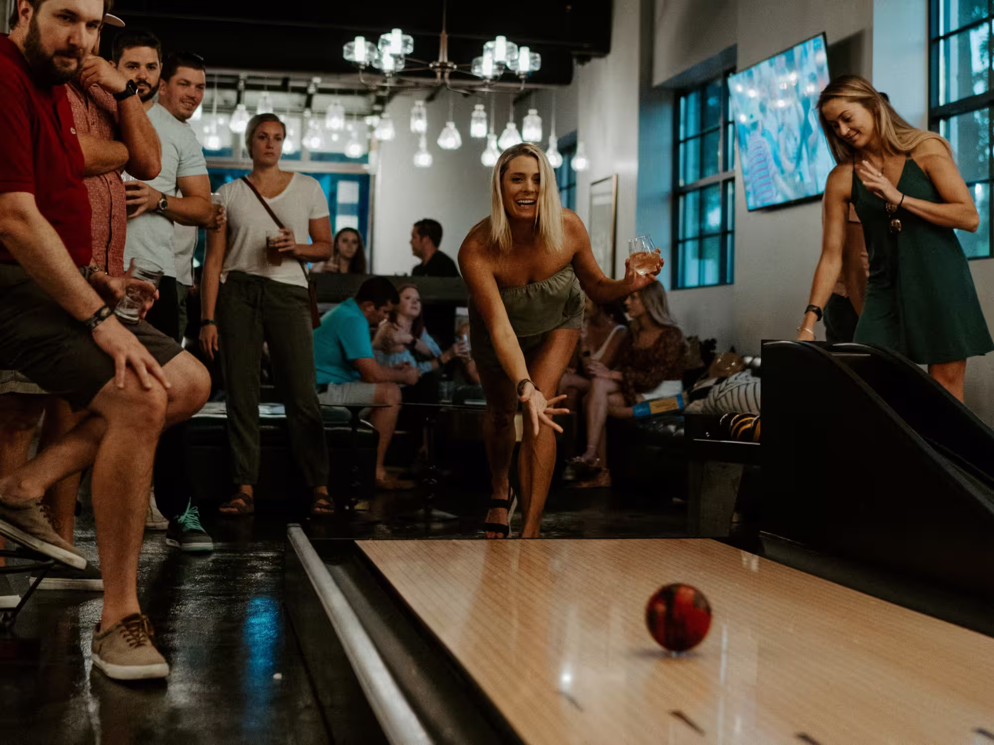 A group of young adults socializing at an indoor bowling alley where a blonde woman is releasing a bowling ball down the lane while holding a drink, and others watch and chat in the background.