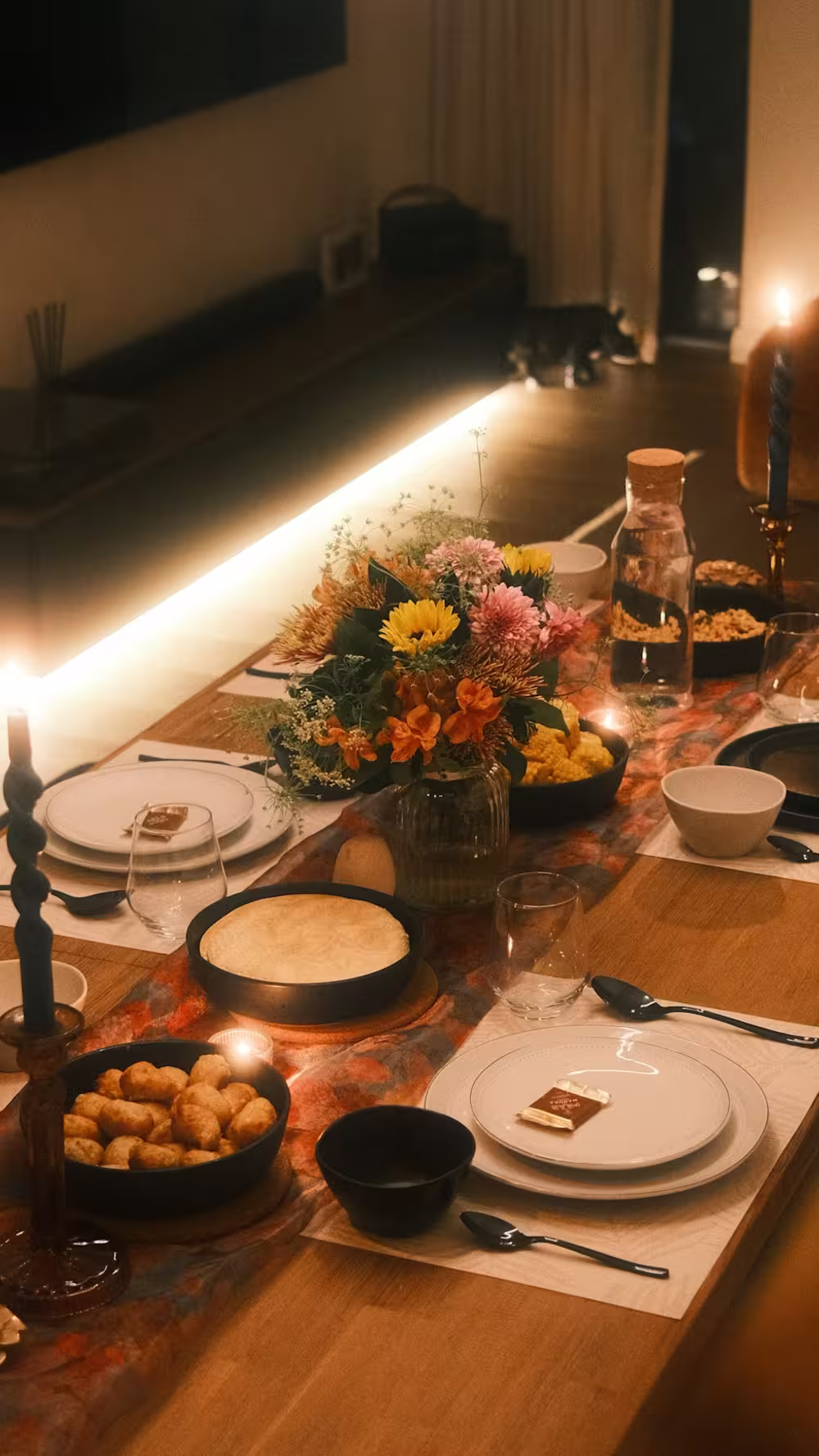 Cozy dining table set for a meal with white plates, black utensils, bowls, and glasses on wooden table with a colorful floral centerpiece, candles, a water bottle, and bowls of food in warm ambient lighting.