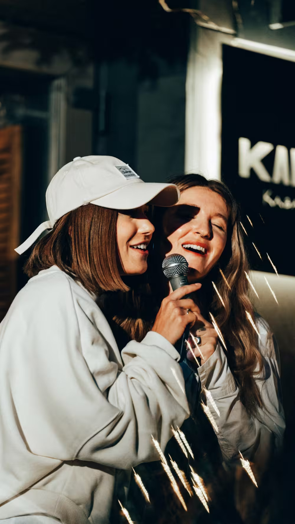 Two young women smiling and singing into a microphone, one wearing a white cap and light-colored sweatshirt, with warm lighting and a dark background.