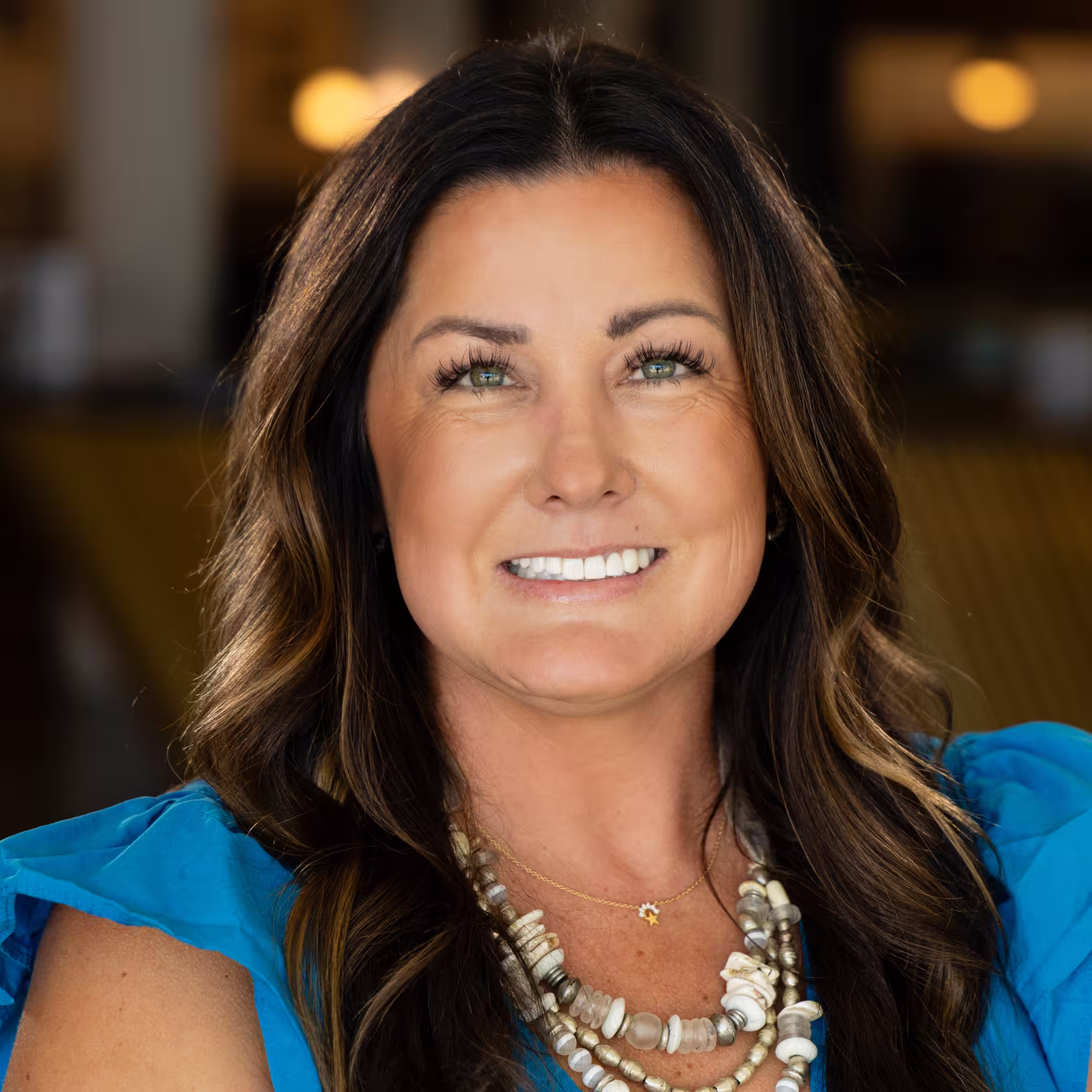 Portrait of a smiling woman with long dark hair wearing a blue top and layered beaded necklaces, against a softly blurred warm background.