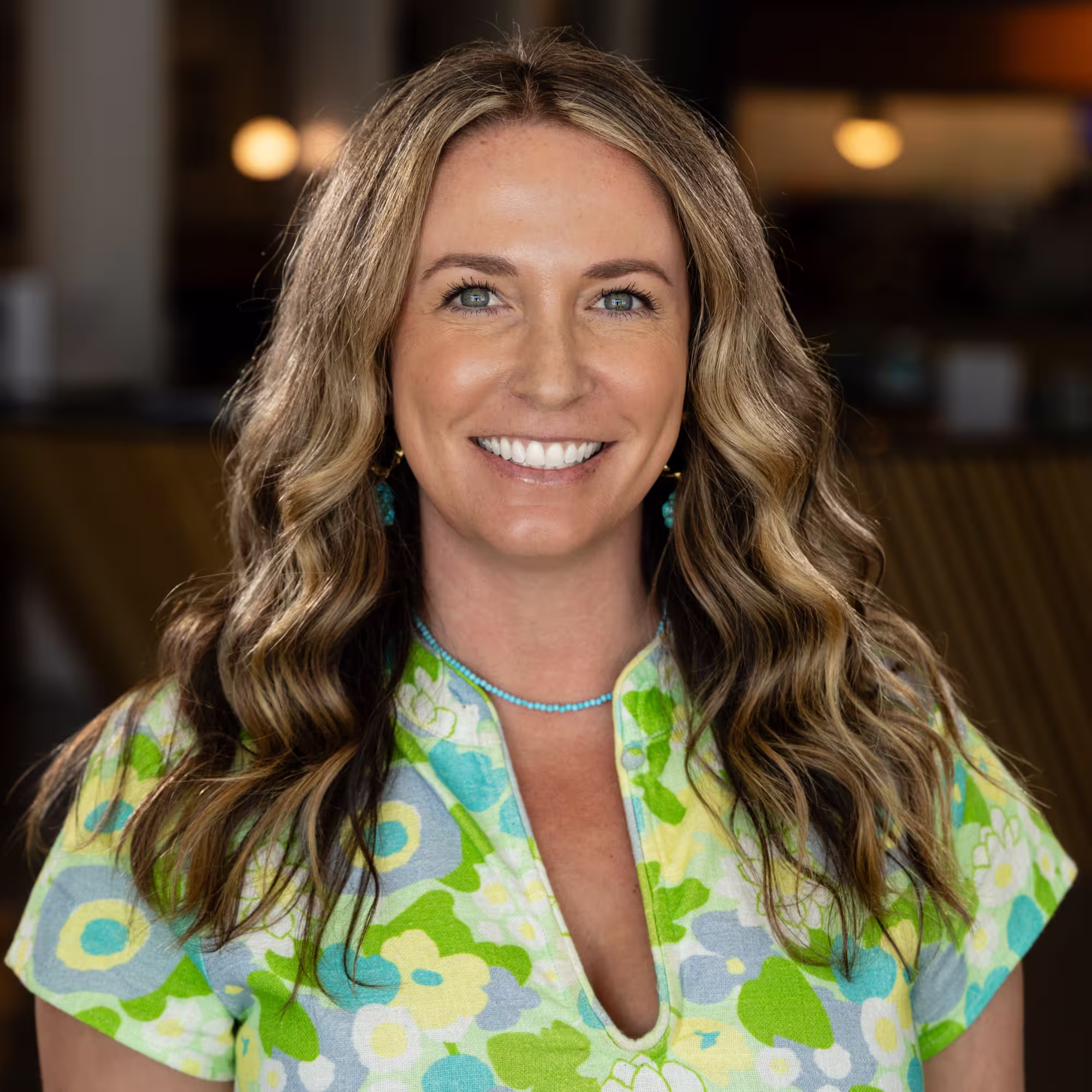Smiling woman with long wavy blonde hair and blue eyes wearing a colorful floral patterned top and turquoise jewelry.