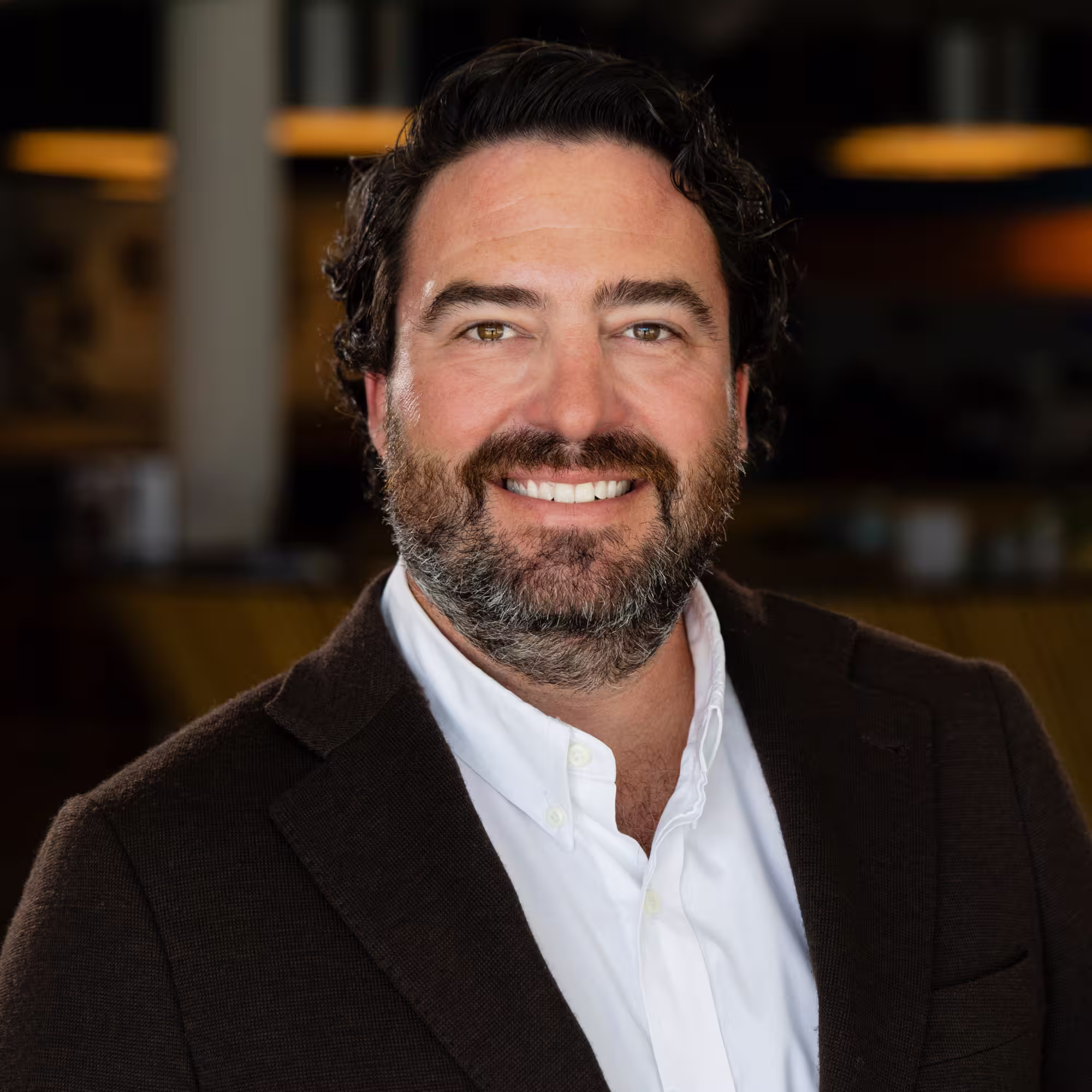 Smiling man with dark curly hair and beard, wearing a white shirt and dark blazer, standing indoors with blurred background lighting.