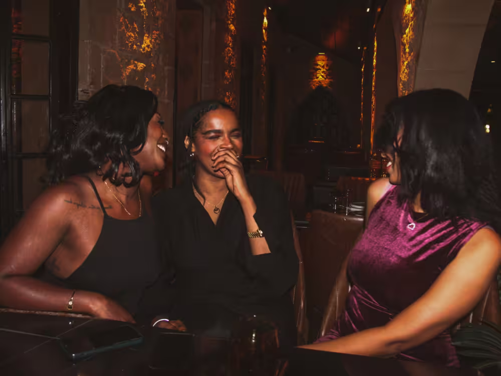 Three women sitting closely together indoors, laughing and enjoying each other's company in a warmly lit setting with textured walls.