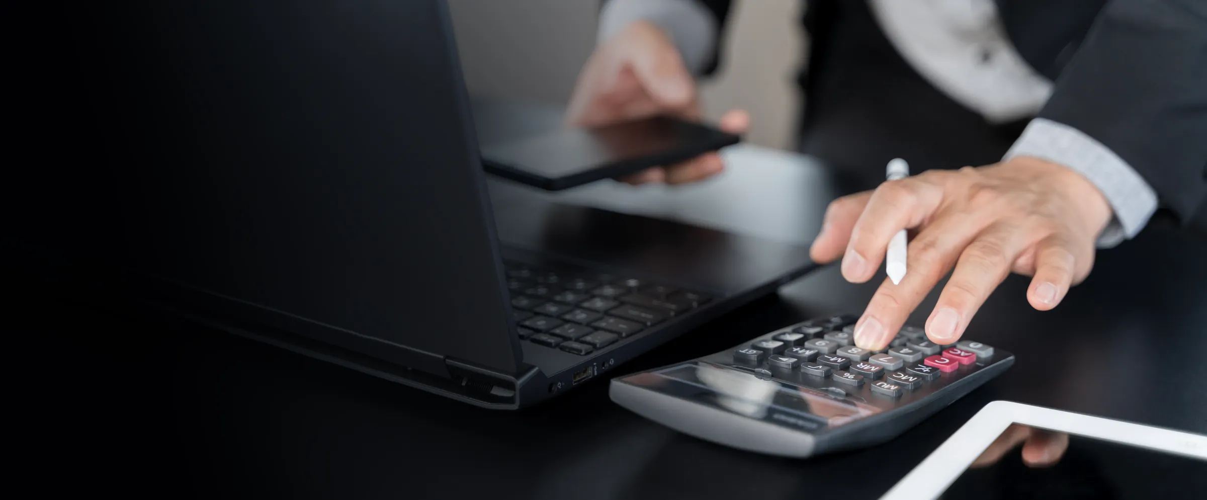 Two professionals at a desk reviewing finances with a calculator, laptop, phone and tablet