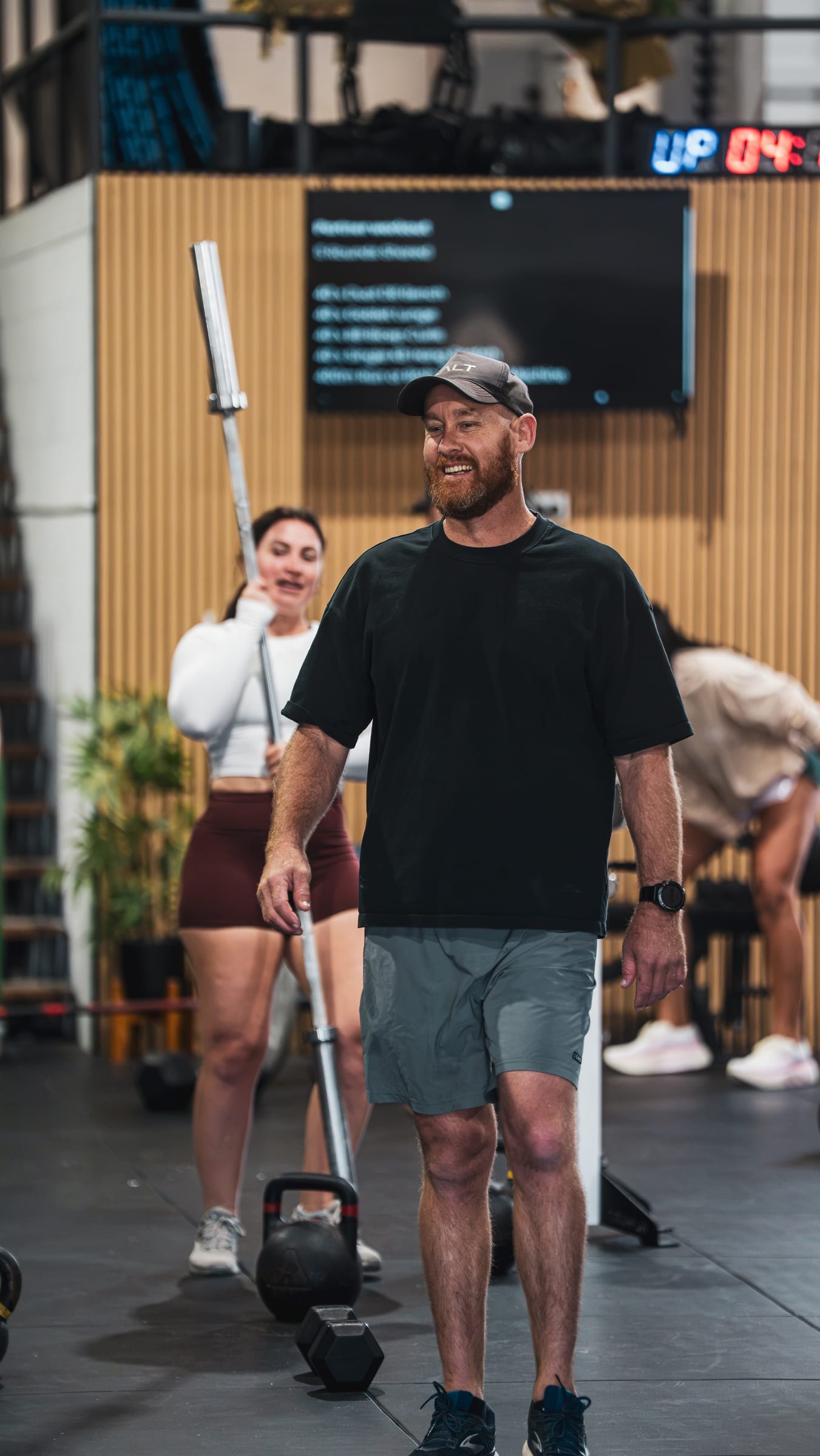 Smiling coach in a black t-shirt and cap walking through the gym with a barbell, a member behind him
