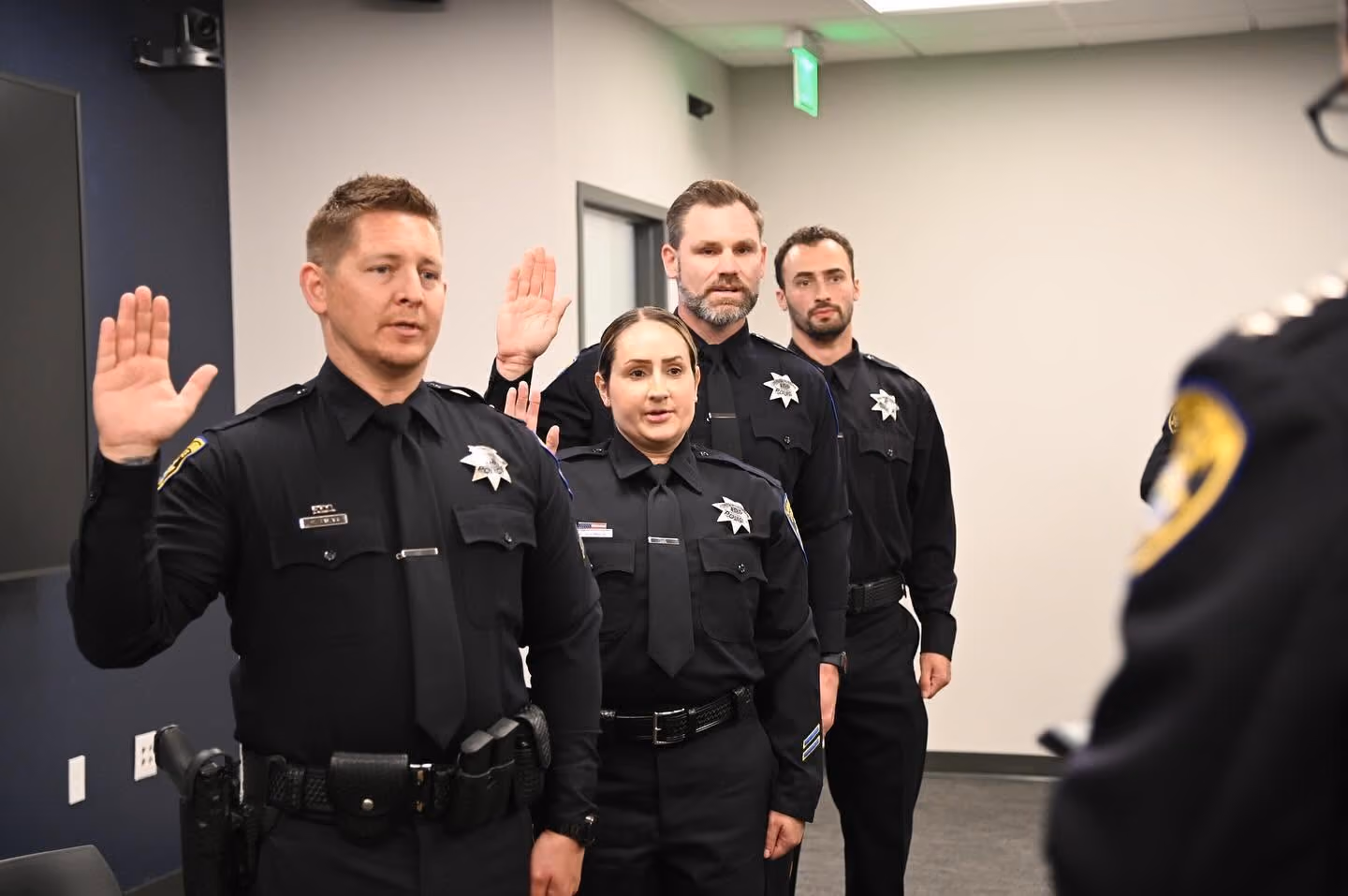 Four police officers in uniform stand indoors raising their right hands, appearing to take an oath or be sworn in.