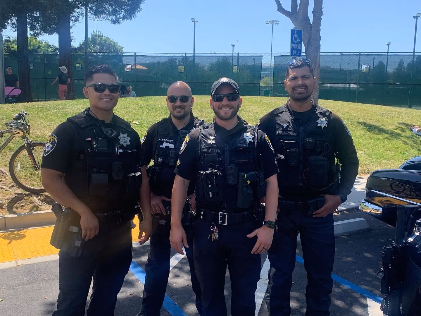 Four male police officers in uniform standing and smiling outdoors on a sunny day with grass and a fence in the background.