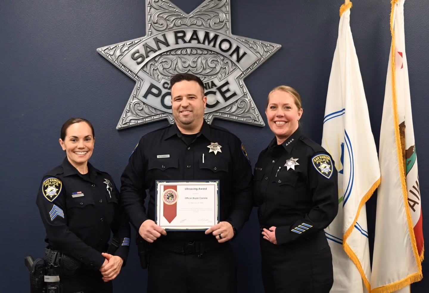 Three police officers from San Ramon Police Department standing in front of a large police badge emblem; the officer in the center holds a framed Lifesaving Award certificate and all are smiling.